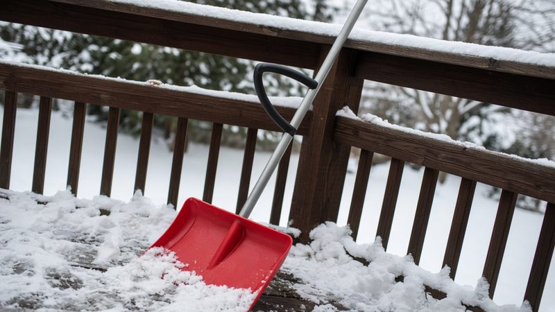 A red snow shovel with a second curved handle leaning on a snowy deck railing, in Thornton, Colorado